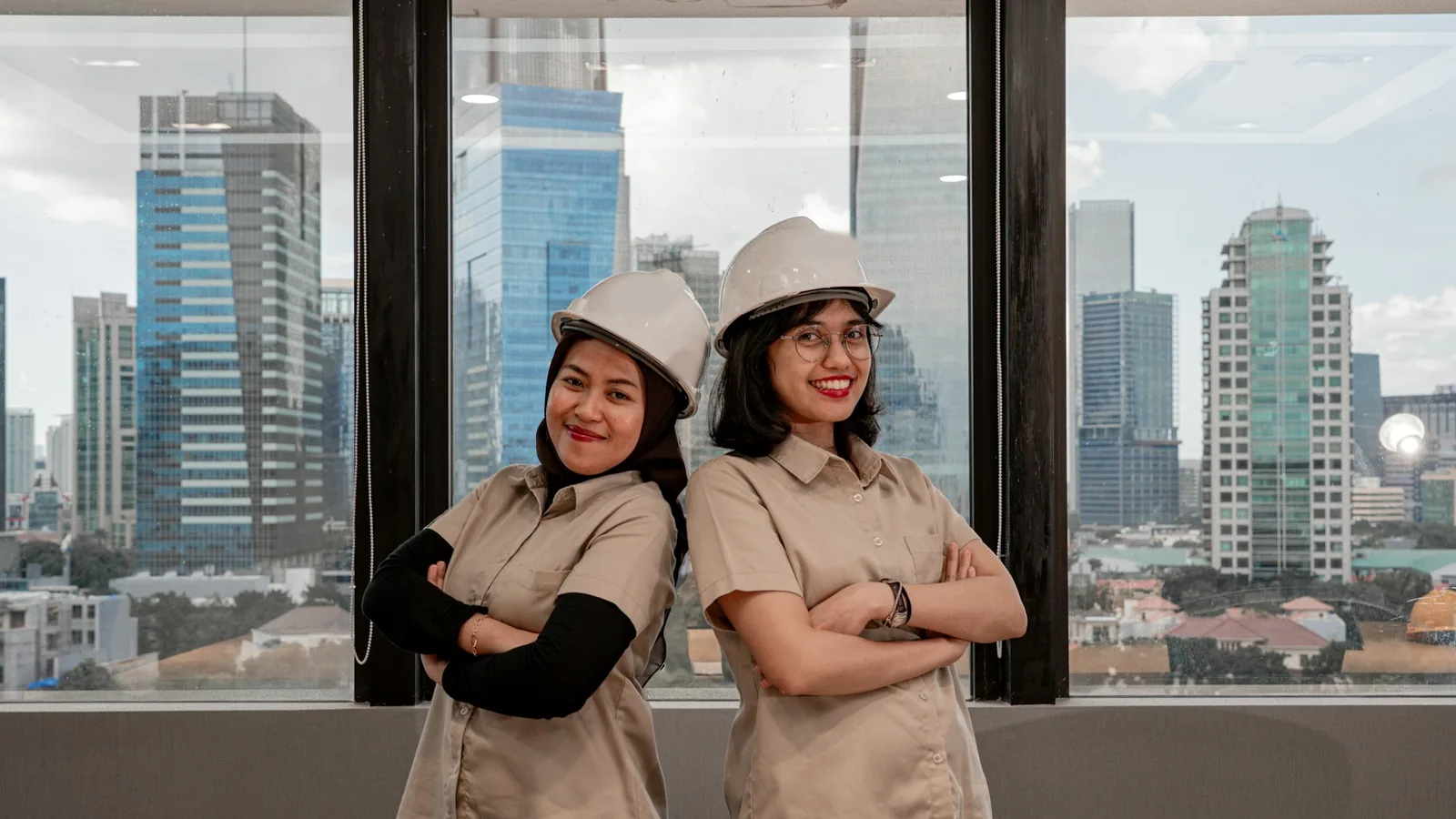 Foto dua pekerja perempuan Indonesia berhelm proyek dan berseragam kerja, tersenyum di depan jendela gedung tinggi kota.