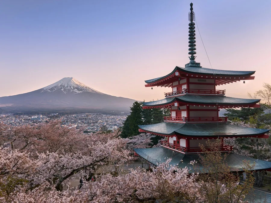 Foto pagoda merah lima tingkat di antara pohon sakura yang mekar, dengan Gunung Fuji bersalju di kejauhan.