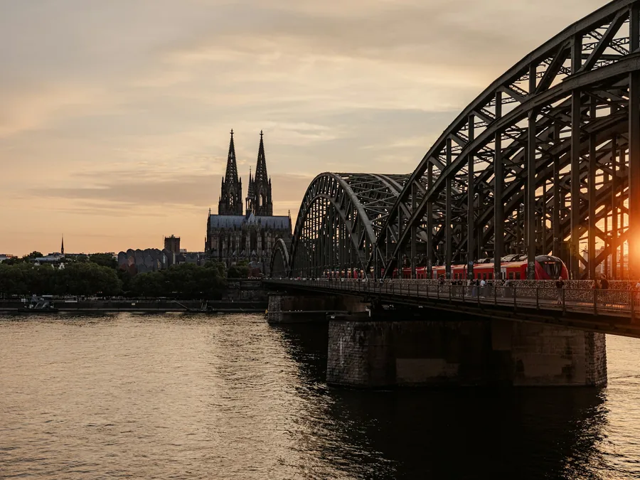 Foto Katedral Koln dengan dua menara tinggi dan jembatan baja melengkung di atas Sungai Rhein saat matahari terbenam.