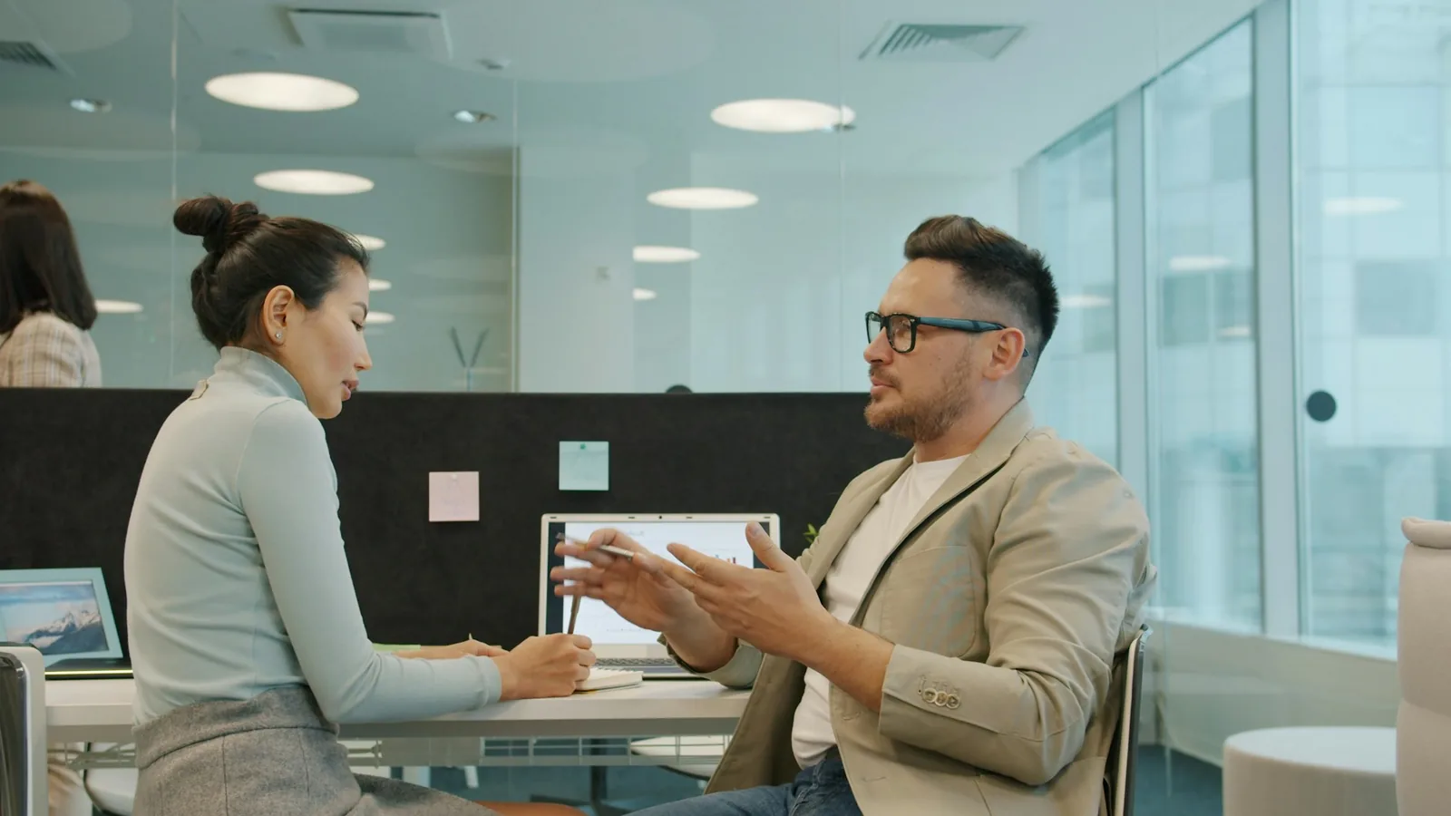 Foto dua profesional berdiskusi di meja kantor modern berdinding kaca, seorang perempuan dan seorang laki-laki berkacamata.