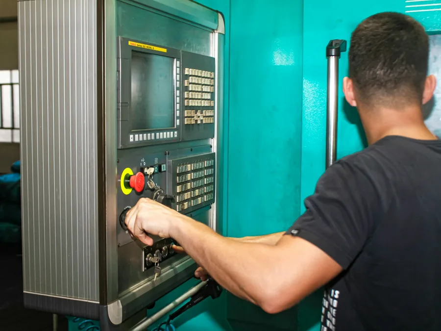 Foto operator berkaos hitam menekan tombol panel kendali mesin CNC berwarna hijau toska.